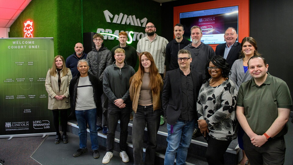 A group of seventeen people stands smiling in a tech-themed room, with vibrant green branding and a digital display. "Welcome Cohort One!" banner is visible.