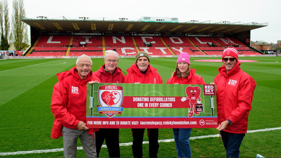 Five people in red jackets hold a banner promoting defibrillator donations at a soccer stadium.