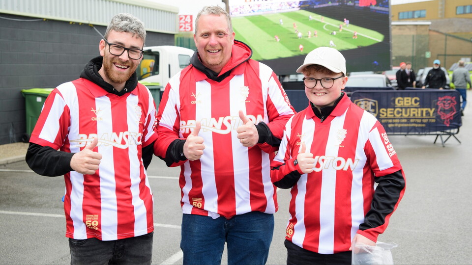 Three people in red and white striped shirts give thumbs-up, smiling openly. A large screen shows a football match in the background.