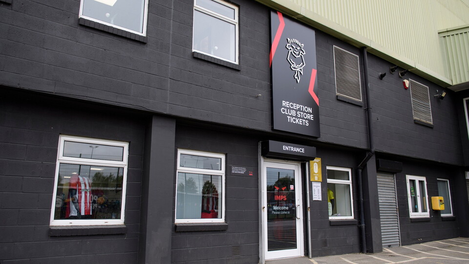 Exterior of a club store with a black facade and large windows displaying sports merchandise. Bold signage reads "Reception, Club Store, Tickets" above the entrance.