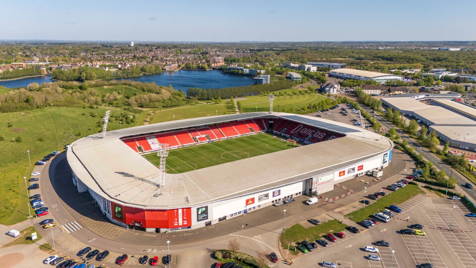 Aerial view of a football stadium with a green pitch and empty red seats. Surrounding areas include parking centres, roads, and lush greenery.