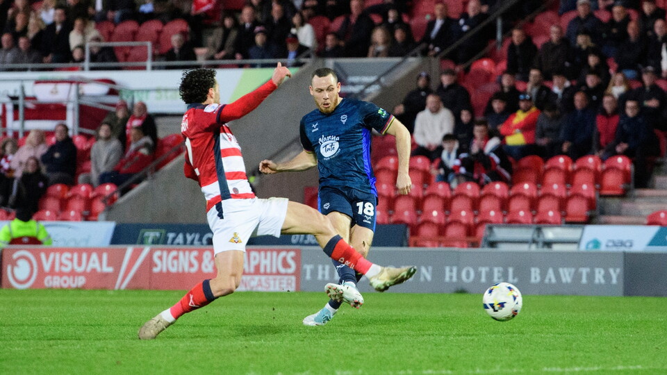 A football player in a blue kit kicks the ball past an opponent in a red and white kit on a green field. The stadium seats are filled with fans.