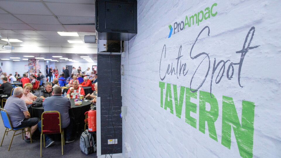 A lively indoor gathering at "Centre Spot Tavern," seen on a white brick wall. Attendees, seated at tables, engage in conversations in a bright, casual setting.