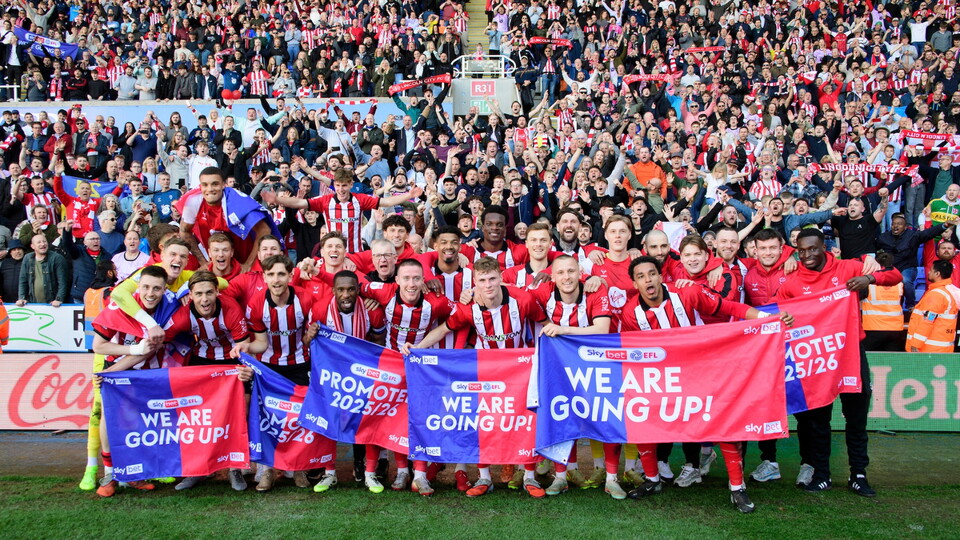 A jubilant football team in red and white striped shirts celebrates promotion with fans. Players hold banners saying, 'We Are Going Up!' Fans cheer wildly.