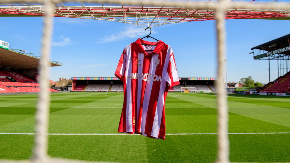 A red and white striped football shirt hangs on a goalpost against a sunny, empty football stadium. The green field and stands are visible in the background.