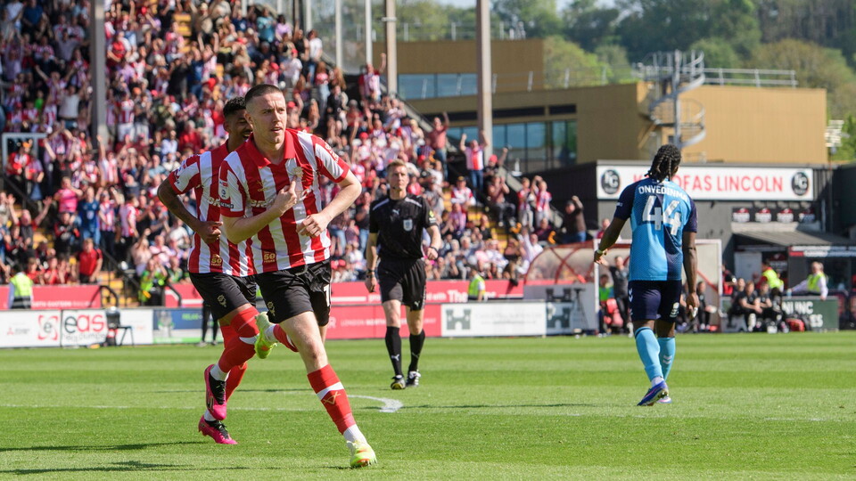 A football player in a red and white striped shirt celebrates on the field, audiences cheer in the stands under a clear blue sky, exuding excitement.