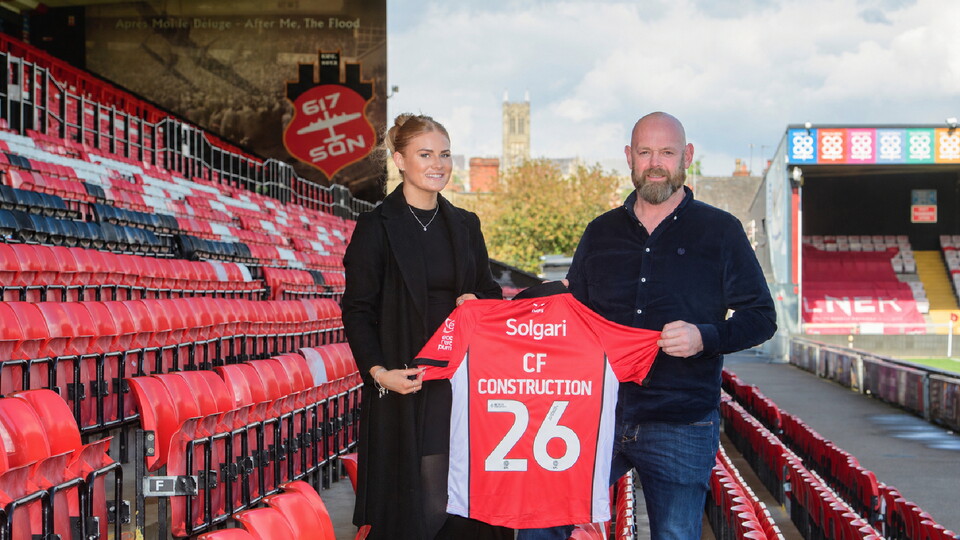 A man and woman stand in a stadium holding a red football jersey labeled 'CF Construction 26'. 