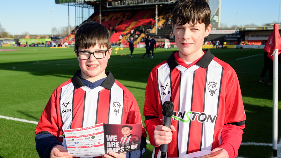 Two boys in red and white striped football jerseys stand on a football field, smiling. One holds a teamsheet and the other, a microphone. Stadium seats in background.