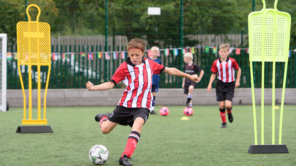 A young boy in a red and white striped top concentrates intensely as he kicks a football on a field. Other kids and training mannequins are in the background.