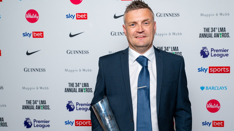 Michael Skubala holds a trophy while stood in front of a sponsors' backdrop