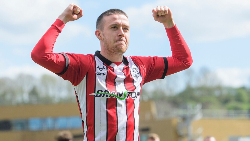 A football player in a red and white striped jersey celebrates with arms raised, displaying determination. The background shows a blurred stadium setting.