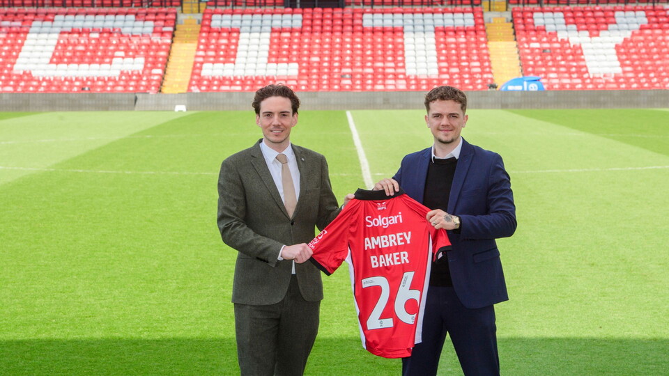 Two men stand on a football field holding a red jersey with "AMBREY BAKER" and number 26. Stadium seats spell "CITY" in white. Both smiling formally.