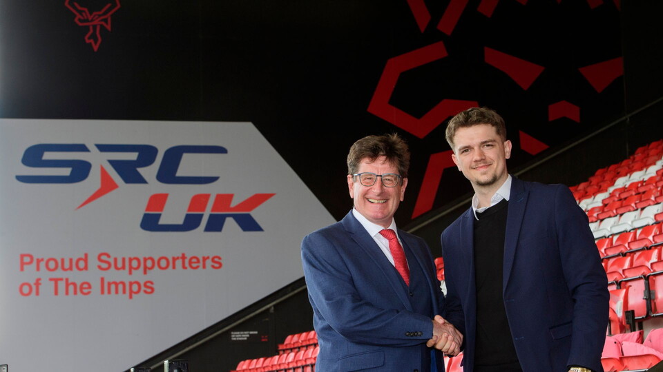 Two men shake hands in a stadium with red seats. "SRC UK Proud Supporters of The Imps" is visible on a sign. The mood is positive and professional.