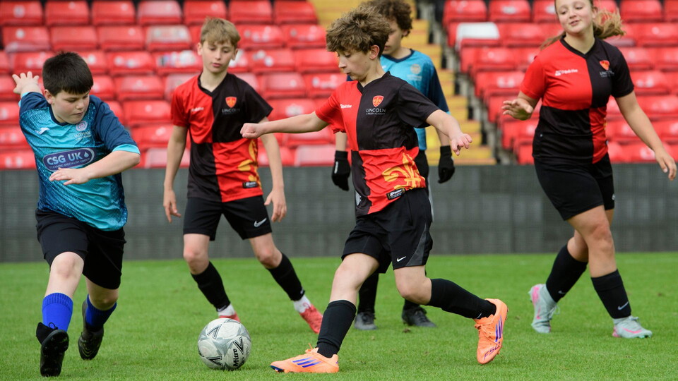 Children playing soccer on a football pitch, wearing red and black or blue jerseys. The focus is on a boy in red dribbling the ball, with others watching.