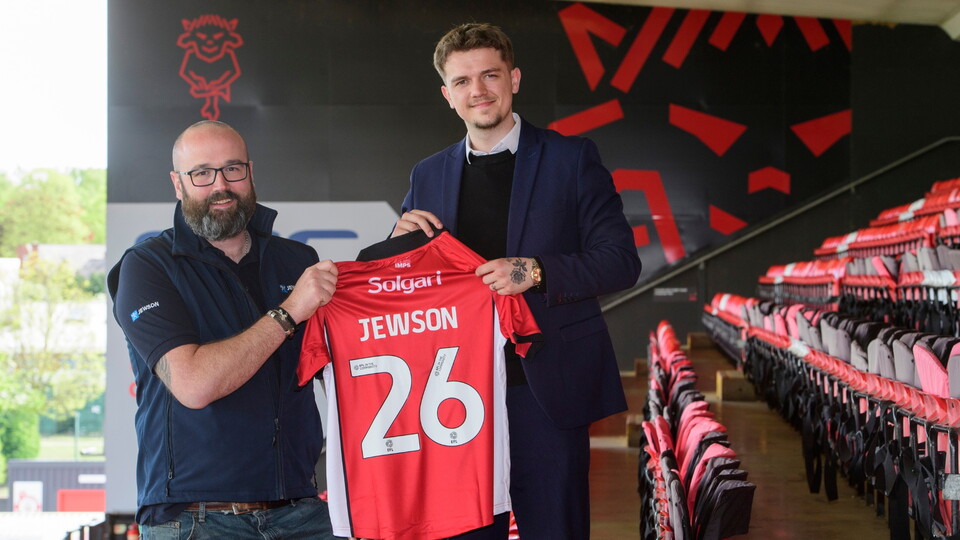 Two men in a stadium stand, smiling while holding a red and white football shirt with 'Jewson 26' printed on it.