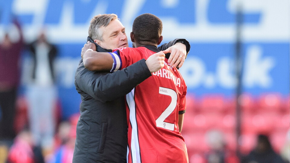 A coach embraces a football player in a red jersey with number 2, celebrating joyfully on the field. The background shows blurred spectators and stadium elements.