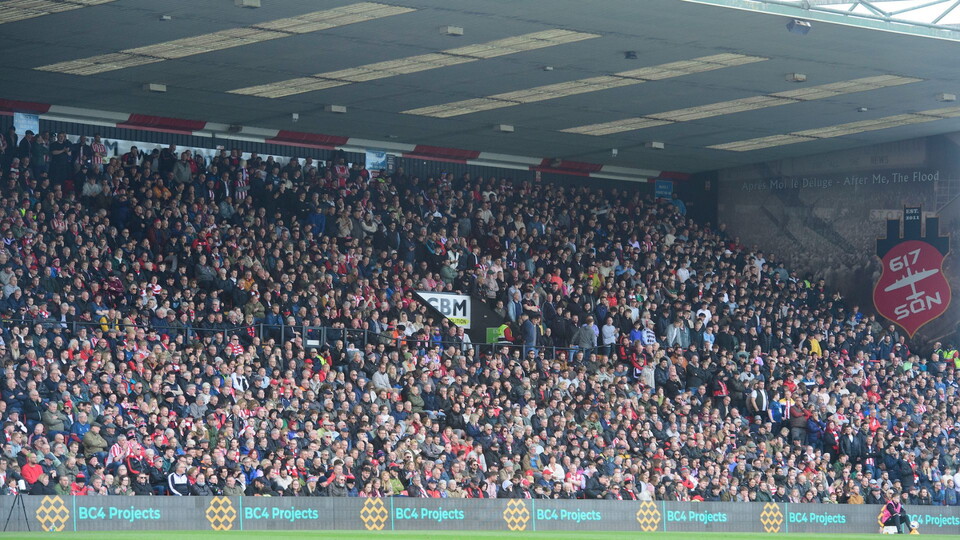 Packed football stadium with cheering fans, many wearing red scarves and jerseys, creating a vibrant and energetic atmosphere. Advertising banners line the field.