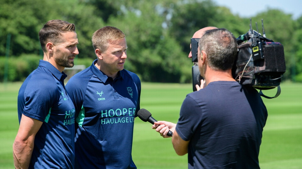 Two men in blue sports attire being interviewed on a lush green field. A cameraman films while a reporter holds a microphone. 