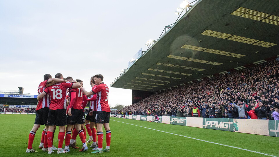 A football team in red and black kits celebrate a goal, huddled near a large, cheering crowd under a covered stadium stand.