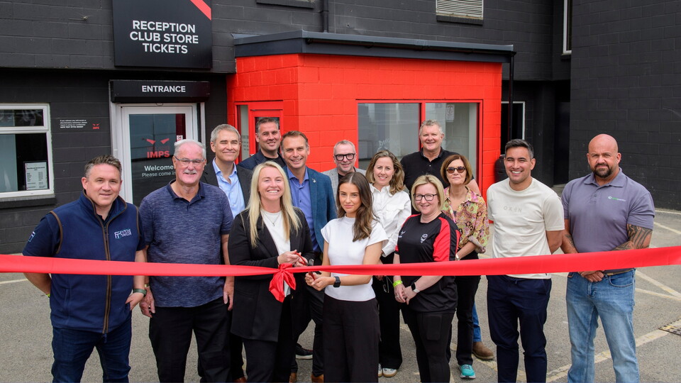 A group of smiling people stand in front of a red and black building entrance, holding a large red ribbon about to be cut, signifying an opening ceremony.