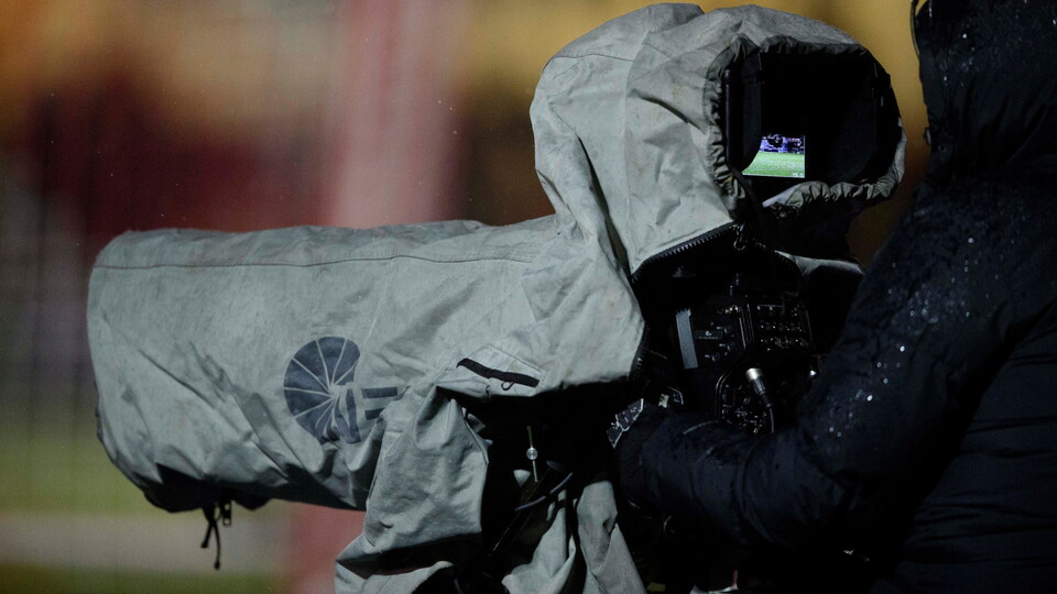 A camera operator wearing a dark jacket films with a large video camera covered by a protective gray rain cover.