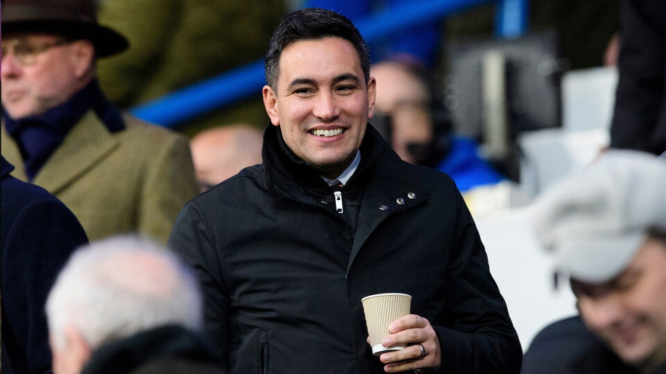 A man in a black jacket smiles warmly while holding a coffee cup in a crowd. 