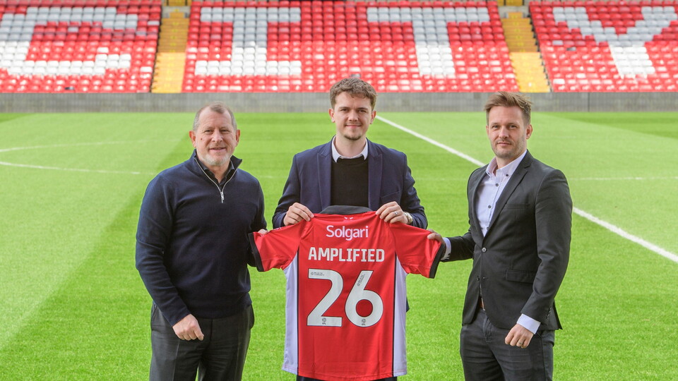 Three men stand on a football field holding a red jersey with 'Amplified' and number 26. The background shows red stadium seats.