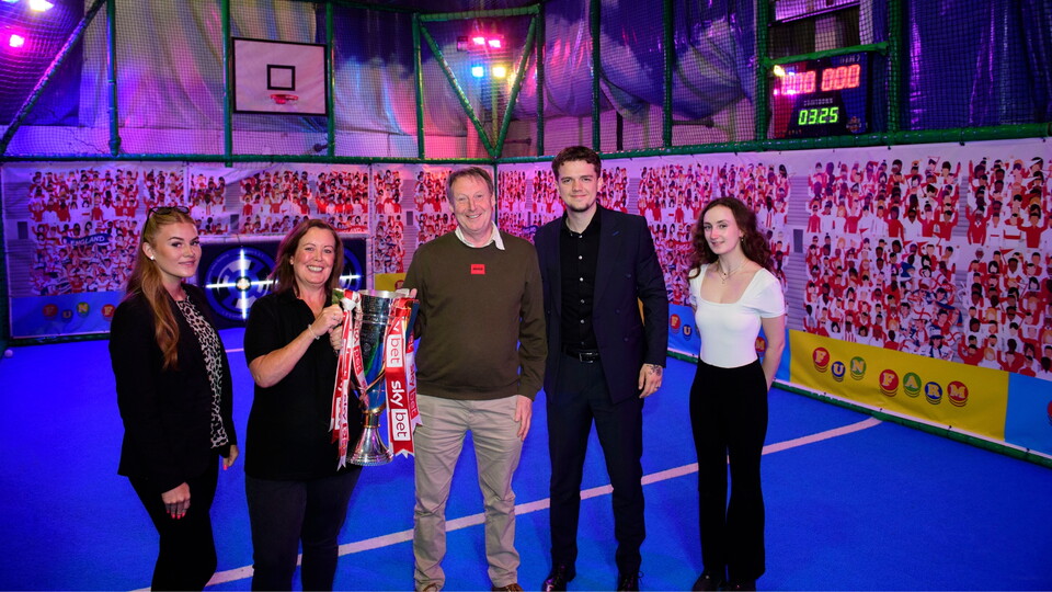 Five people stand on a blue indoor sports court with vivid lighting. They smile while holding a trophy. The backdrop features colorful banners.