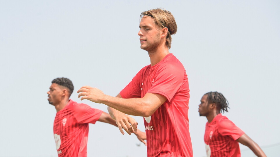 A man in a red training top warming up with two other men in the same attire in the background.