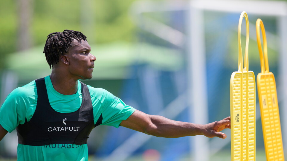 A man in a teal t-shirt points. He is on a football pitch.