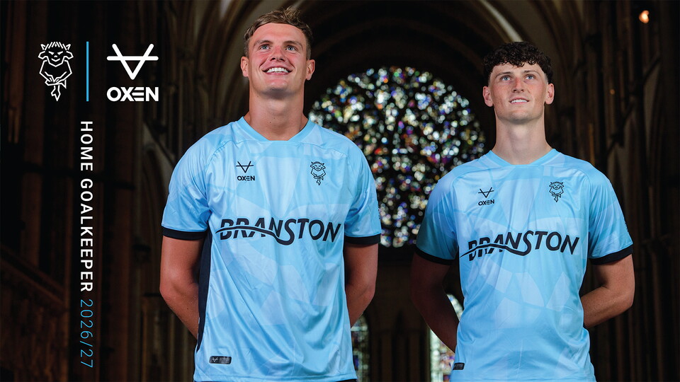 Two men stand in blue goalkeeper shirts posing in front of a stained glass window