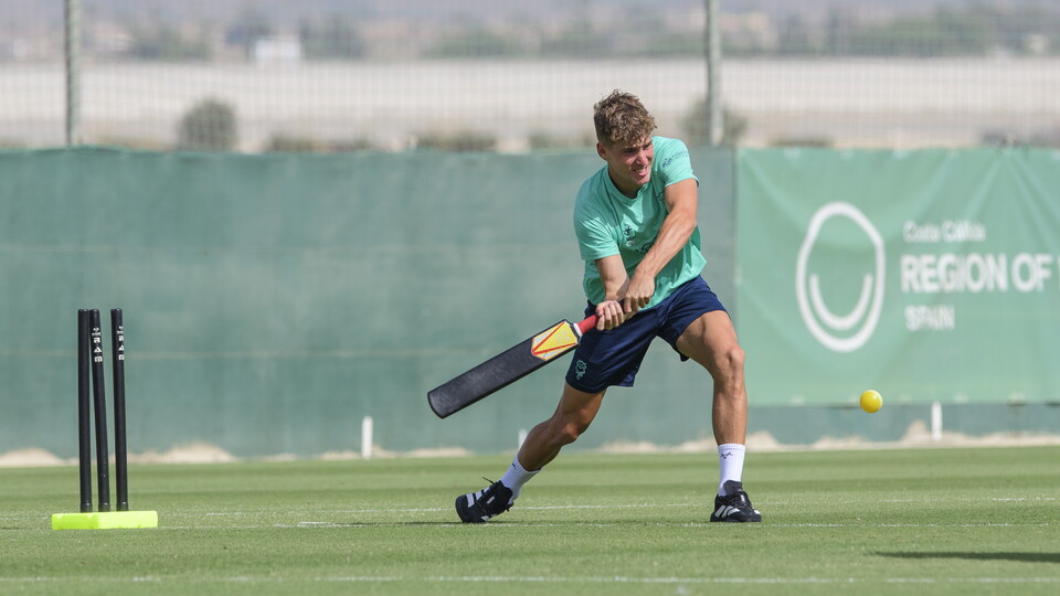 A man in a teal top swings a cricket bat at a ball