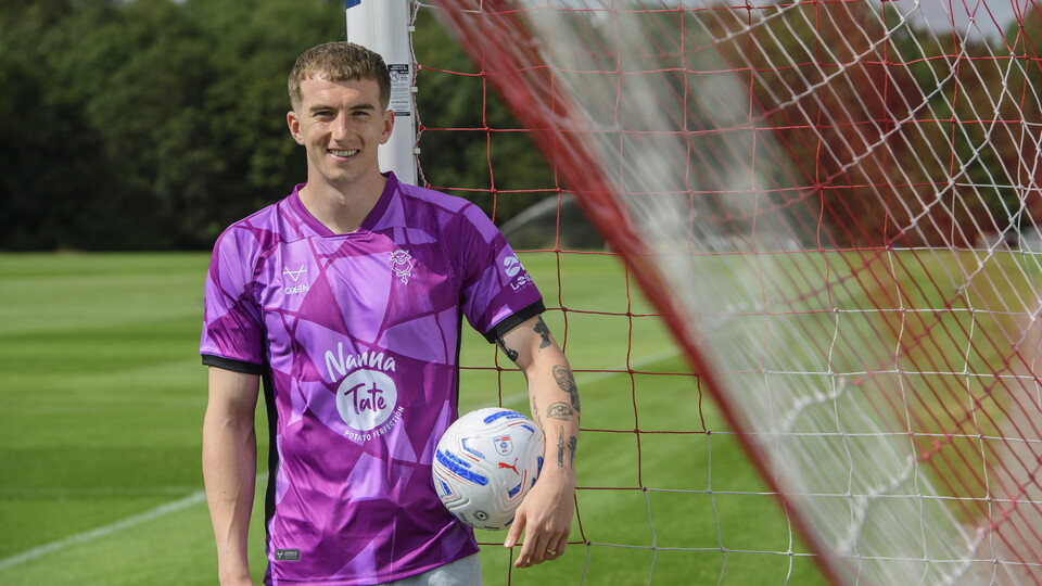 A man wears a purple goalkeeper top and holds a football under an arm.