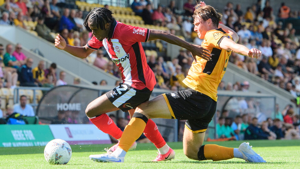A man in a red and white football top shields a football from a man in an amber top.