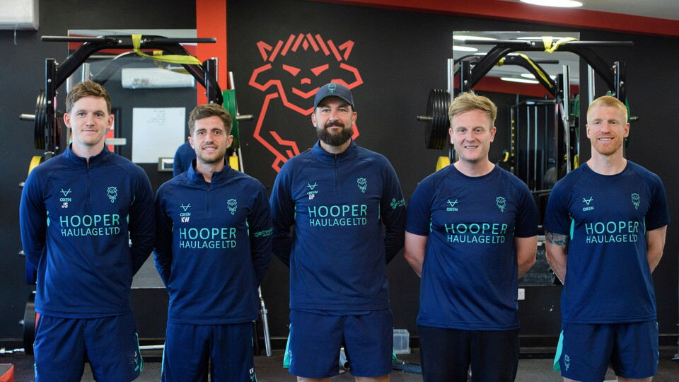 Five men in matching athletic gear stand side by side in a gym setting, smiling. The wall behind them features a stylized red Imp logo.