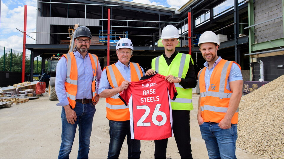 Four men in construction gear and hard hats stand at a building site. They hold a red jersey displaying 'Rase Steels 26' with a new structure being erected in the background. 