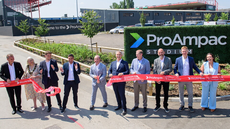 A group of people stands in front of a large sign for ProAmpac Stadium, holding a red ribbon during a ribbon-cutting ceremony. They are dressed in formal attire, with a green landscape and buildings visible in the background.