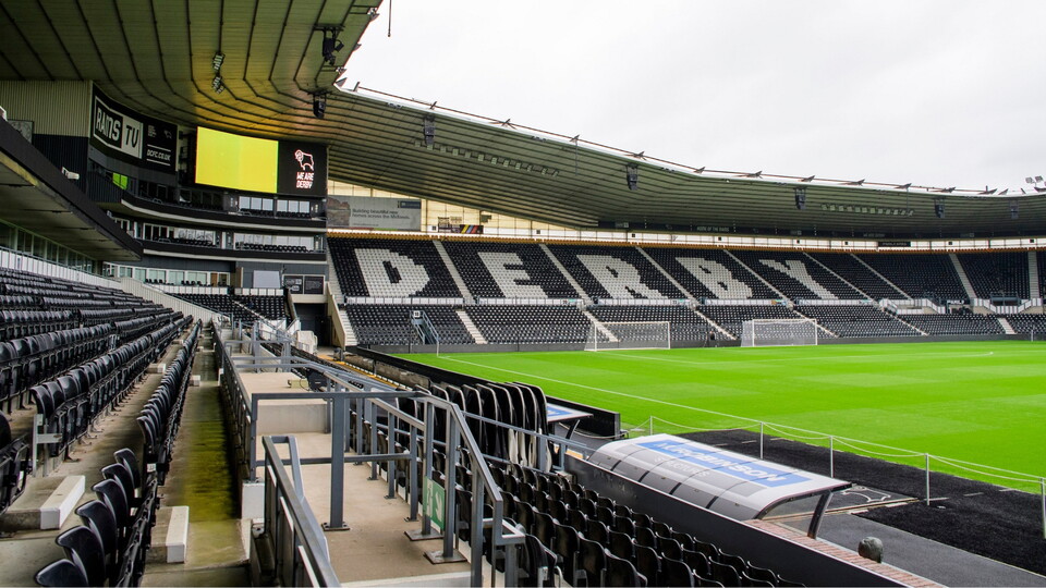 A wide-angle view inside a football stadium showing empty black-and-white seats spelling “DERBY” across the stand, a green pitch, team dugout, large scoreboard, and covered seating areas under an overcast sky.