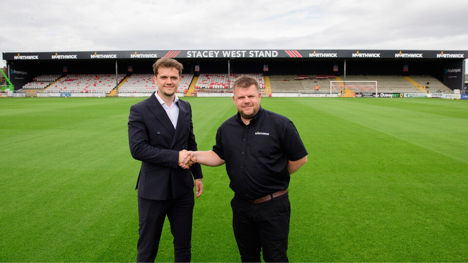 Two men shaking hands on a football pitch with the Stacey West Stand in the background