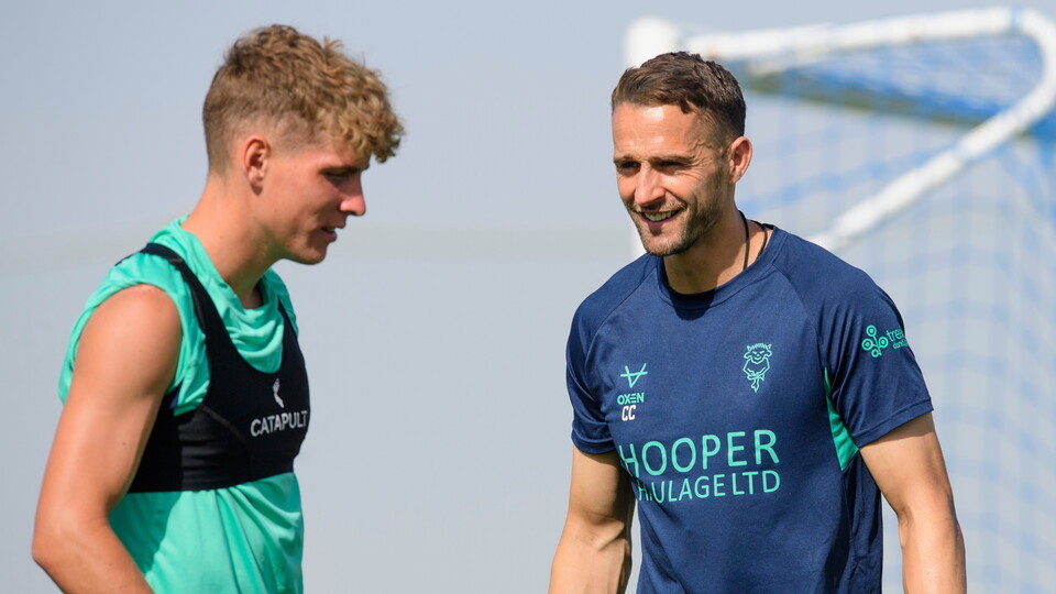 Two men in sportswear are conversing happily on a sunny day, with a football goalpost in the background. 