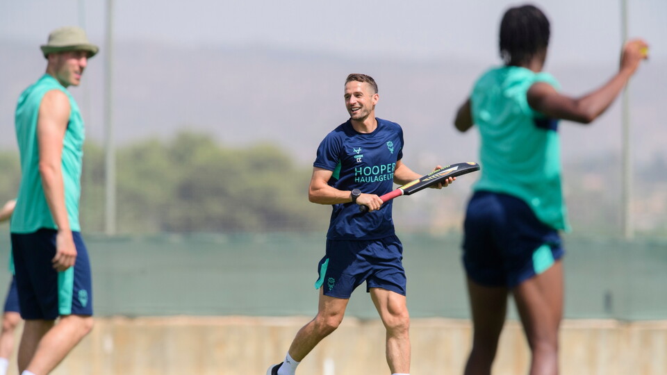 Three men in sportswear are playing with a cricket bat and ball outdoors on a sunny day. One person in the center is smiling, holding a cricket bat.