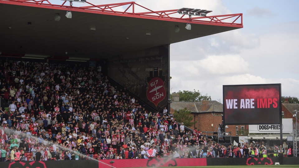 Lincoln City supporters in a stand. There is a big screen which reads WE ARE IMPS