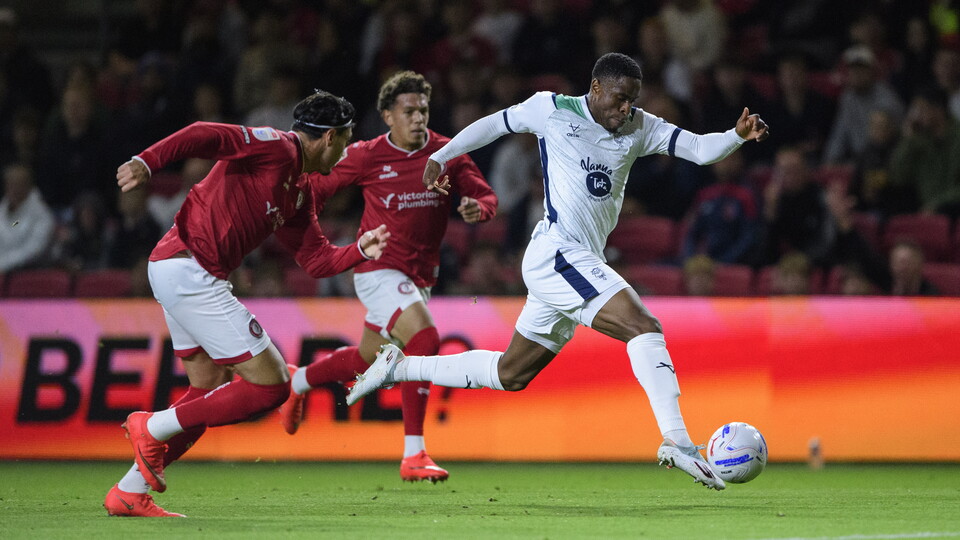 Chieo Ogbene, wearing a white football kit, dribbles with the football away from two players in red and white.