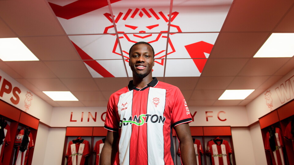Daniel Oyegoke poses in a red and white striped football shirt in front of arranged football shirts in a changing room