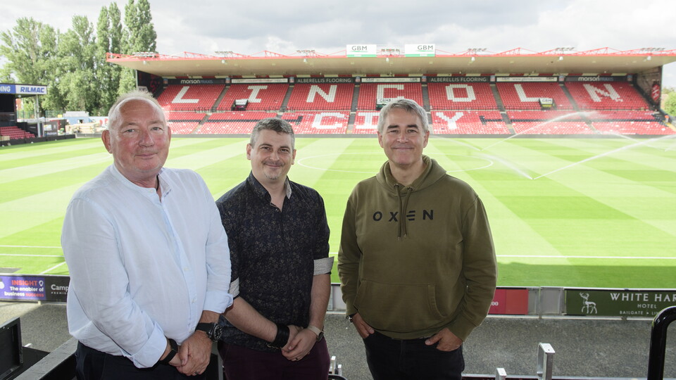 Three men stand in front of a football pitch