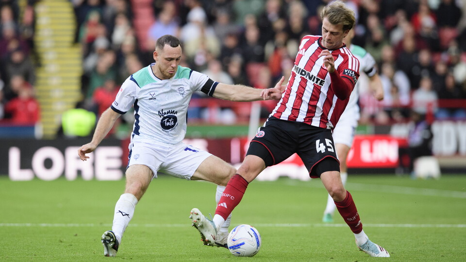 A footballer in white kit challenges a footballer in red and white striped kit who has the ball