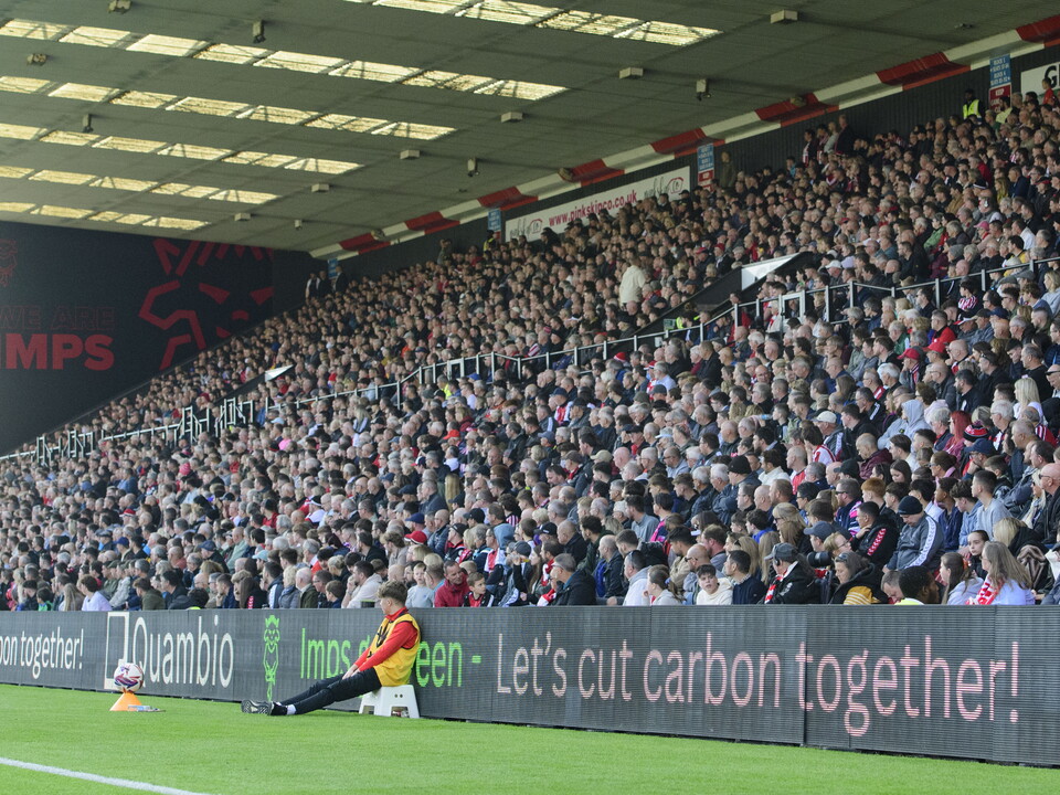 Lincoln City fans watch a game from a packed GBM Stand. At the back of the stand is a mural which reads "We Are Imps".